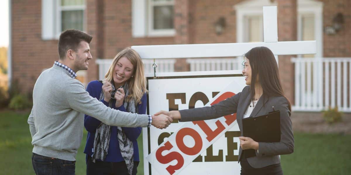 Real estate agent shaking hands with a smiling couple in front of a 'sold' sign.